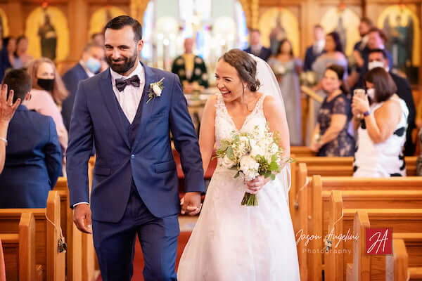 Newlyweds walking u church aisle as guests wearing COVID masks look on