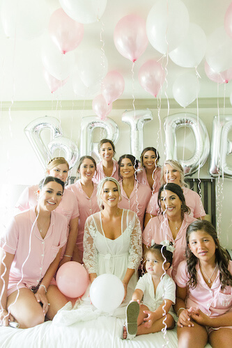 bride on bed surrounded by bridal party in pink robes with silver BRIDE balloons and pink balloons