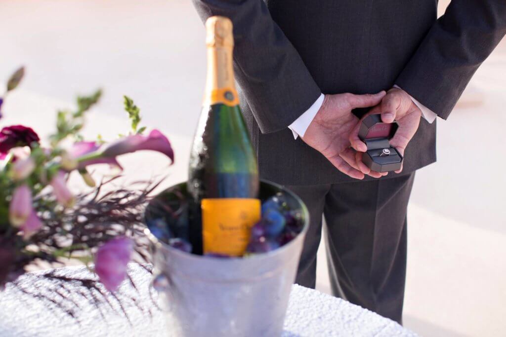 man holding an engagement ring behind his back ready for with a bottle of champagne and flowers next to him for the perfect proposal