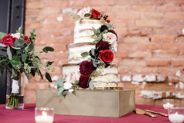 naked wedding cake with burgundy and blush flowers sitting on a wooden box