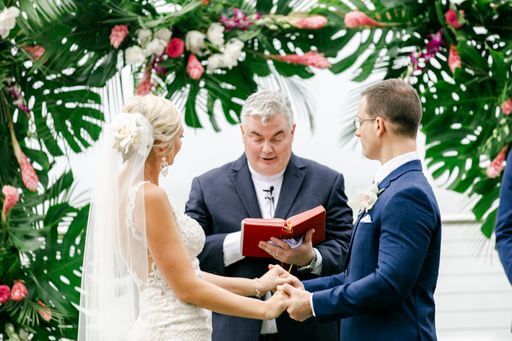 wedding officiant reading to a bride and groom in front of their Hawaiin inspired wedding structure of tropical flowers