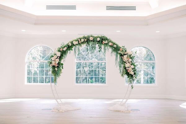 white moon gate wedding arch decorated with lush greenery and pink and white flowers at Harborside Chapel