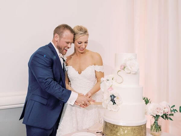 bride and groom cutting elegant four tiered white wedding cake with gold monogram and soft pink flowers