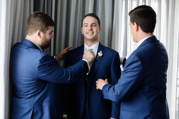 two groomsmen helping groom with finishing touches to his navy blue suit