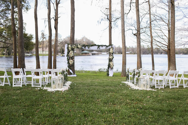 At home waterfront intimate wedding ceremony with white garden chairs, white lanterns and a wedding structure created between two trees