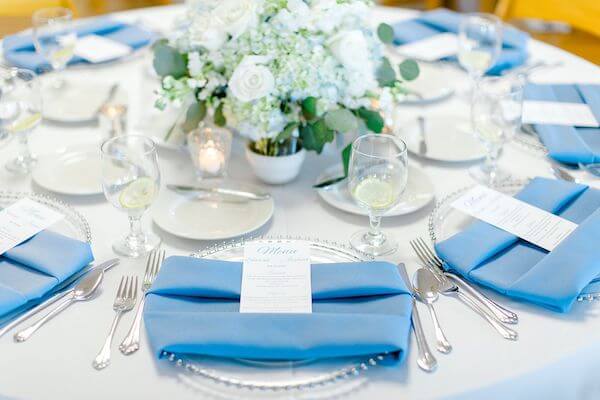 wedding reception at Tampa Bay Watch set with glass charge plates, blue napkins and a custom menu card