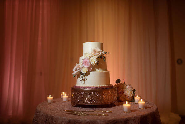 three tiered white wedding cake with pink and white flowers on a table with boho chic pink embroidered lace linen