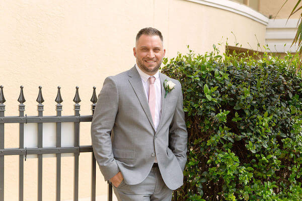 groom wearing a light grey suit pink necktie and white rose boutonniere leaning against a fence