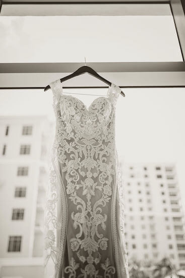 black and white photo of a sheer embroidered lace wedding gown hanging in front of a window