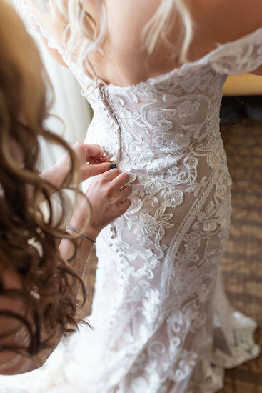 bridesmaid buttoning the back of a form fitting embroidered lace wedding gown