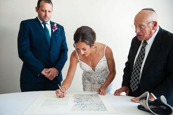 bride signing the ketubah as her groom-to-be and Rabbi look on