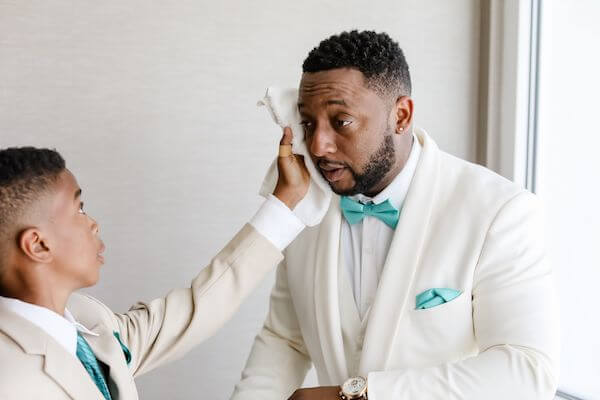 groom in a white tuxedo with seafoam colored tie getting his head cooled by a ring bearer