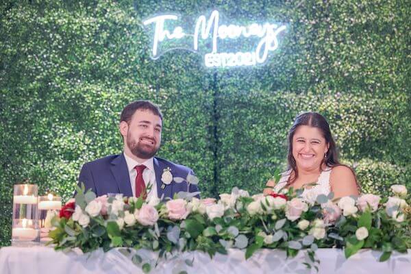 Tampa bride adn groom sitting at thier sweetheart table in front of a hedge wall with a custom neon sign