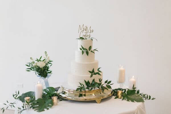 Four tiered white wedding cake surrounded by white flowers, greenery and candles