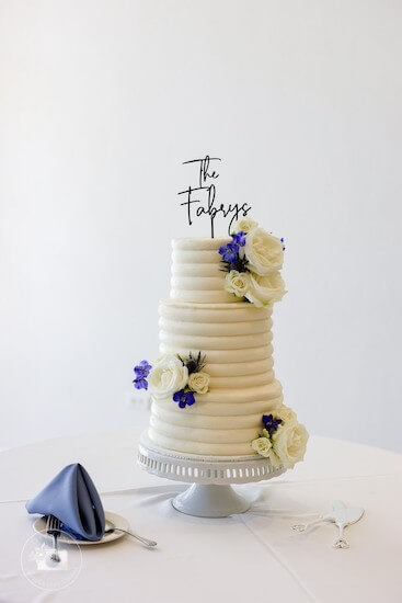 Modern white three-tiered wedding cake with blue flowers and a custom cake topper