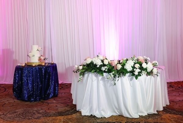 Sweetheart table with lush flowers next to a cake table with blue sequin linen