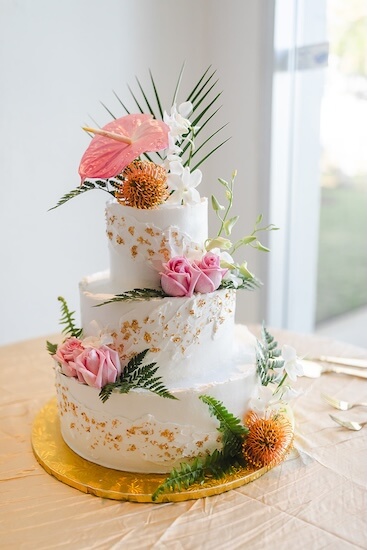 A three tiered white wedding cake with gold leaf and tropical flowers