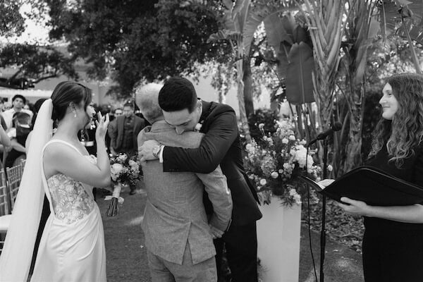 A bride's father hugging the groom as his dauther looks on.